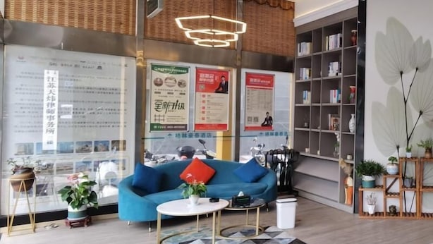 Un área de recepción moderna con un sofá azul, una mesita redonda y una alfombra estampada. Hay carteles en la pared, una estantería y una planta en maceta.