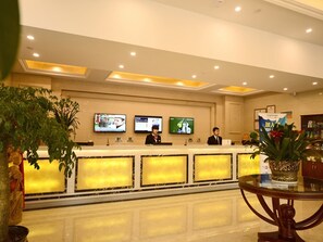 A hotel reception area with a reception desk, two receptionists, three monitors on the wall, and potted plants. - GreenTree Inn Xuancheng Ningguo City East Ningyang Road RT Mart Express Hotel (Xuancheng)