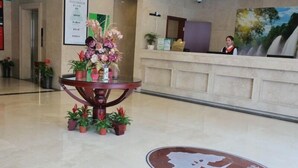 Zona de recepción con mostrador, recepcionista y un cartel de Green Tree Inn. Hay plantas en macetas alrededor de una mesa y un cartel en la pared con texto.