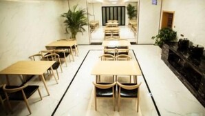 Un salón de conferencias de hotel con varias mesas y sillas, una planta en el centro, y una zona de recepción con mostrador y sillas.