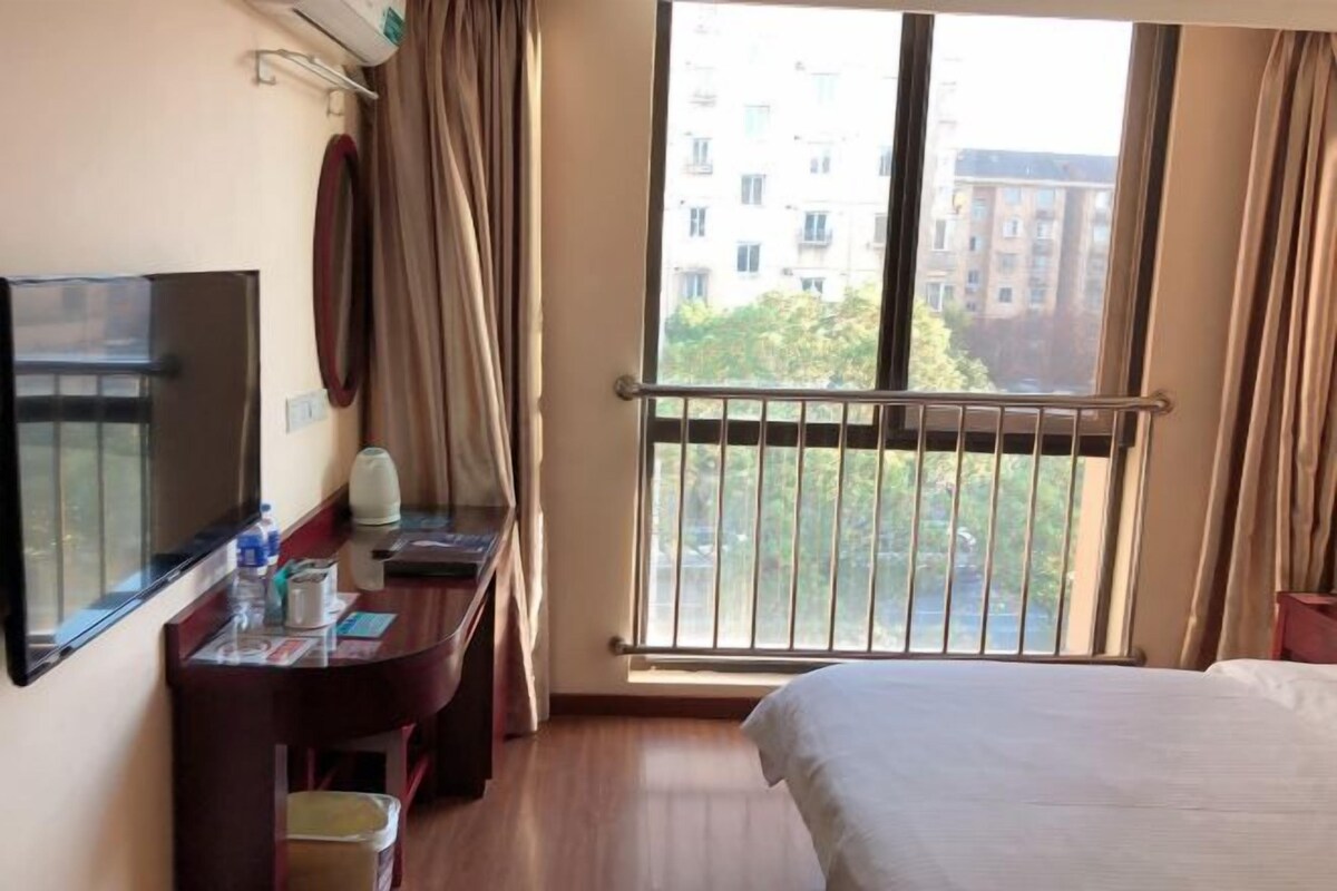 A hotel room with a bed, a desk with a computer, a TV, and a balcony with a view of buildings.