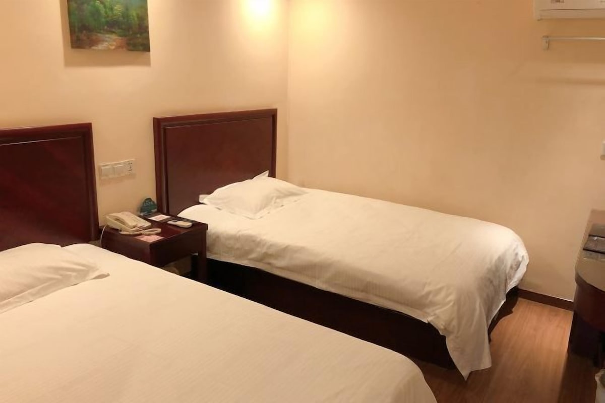 A hotel room with two single beds, a bedside table with a lamp, and a wall-mounted air conditioner.