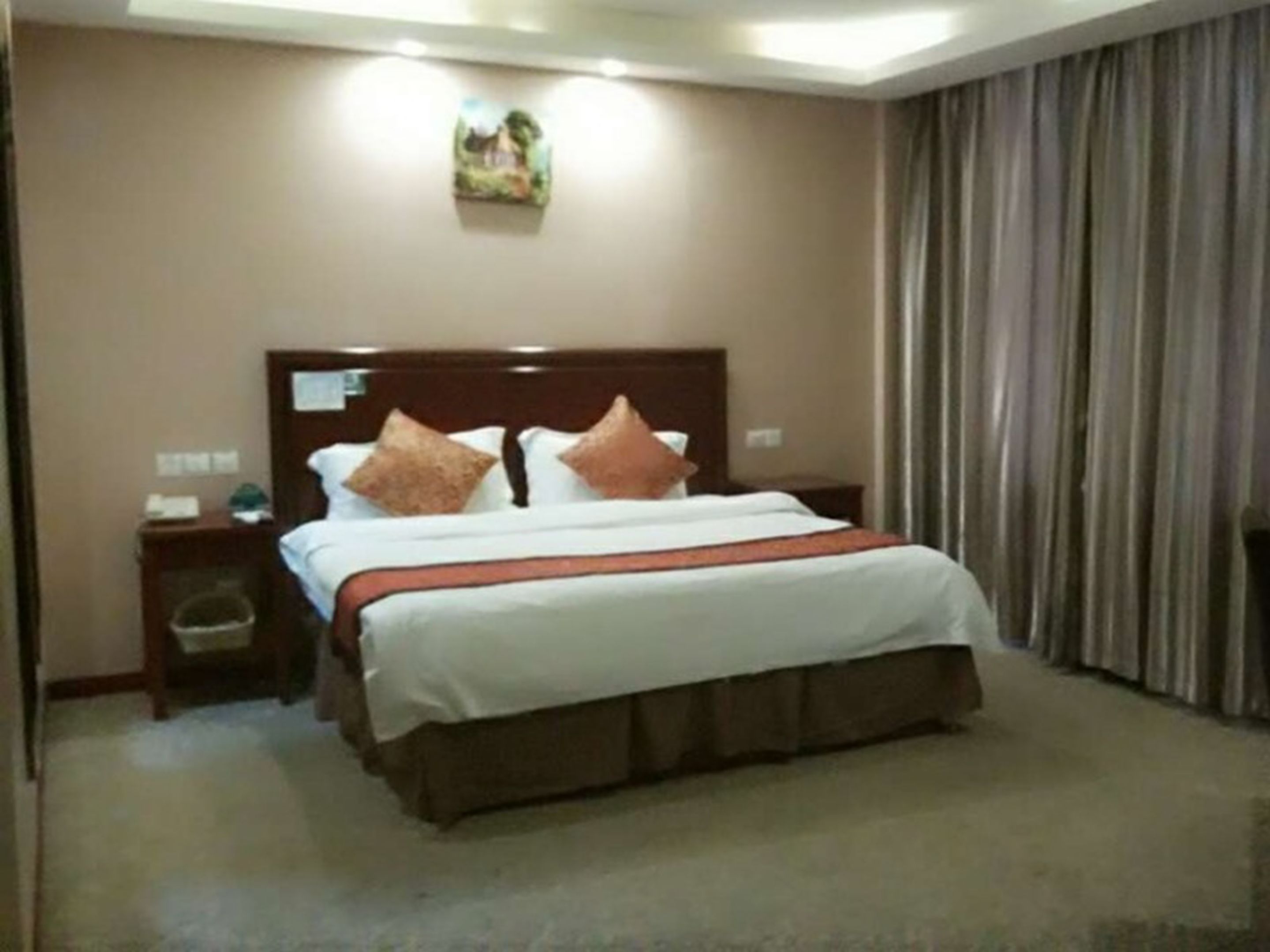 A hotel room with a large bed, a wooden headboard, a nightstand, a trash bin, and a painting on the wall.