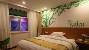A hotel room with a bed, a nightstand, a green chair, a window with a view of lavender fields, and a wall with a tree design and the hotel's name. - Vatica ZaoZhuang TengZhou Railway Station Hotel (Zaozhuang)