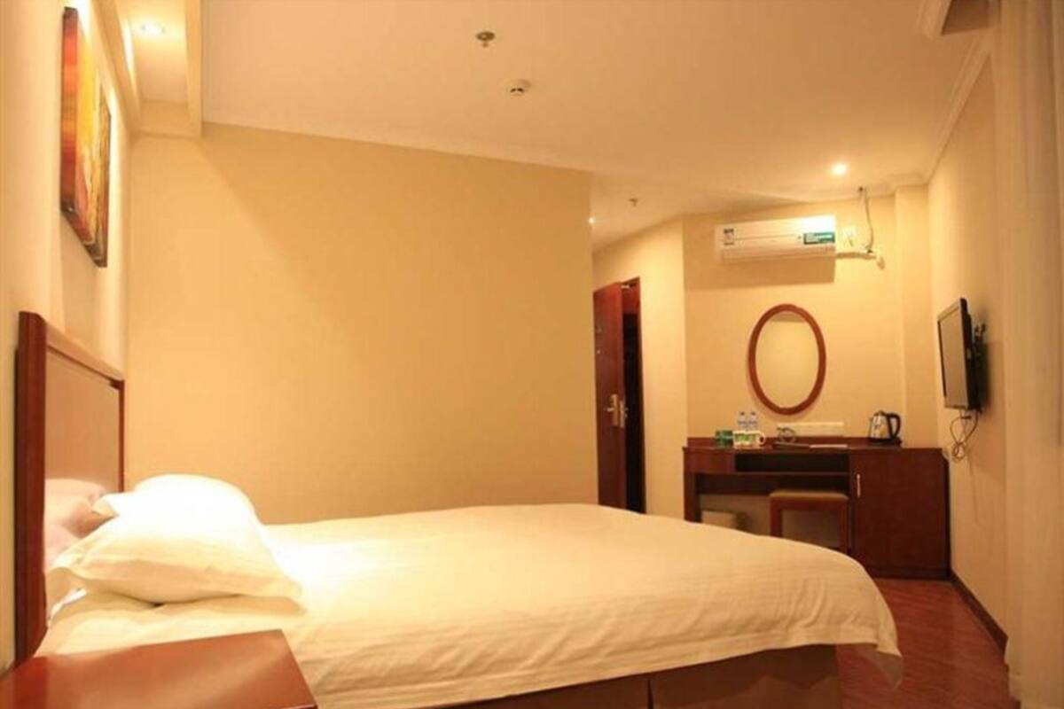 A hotel room with a bed, a desk, a chair, an air conditioning unit, a mirror, and a television.