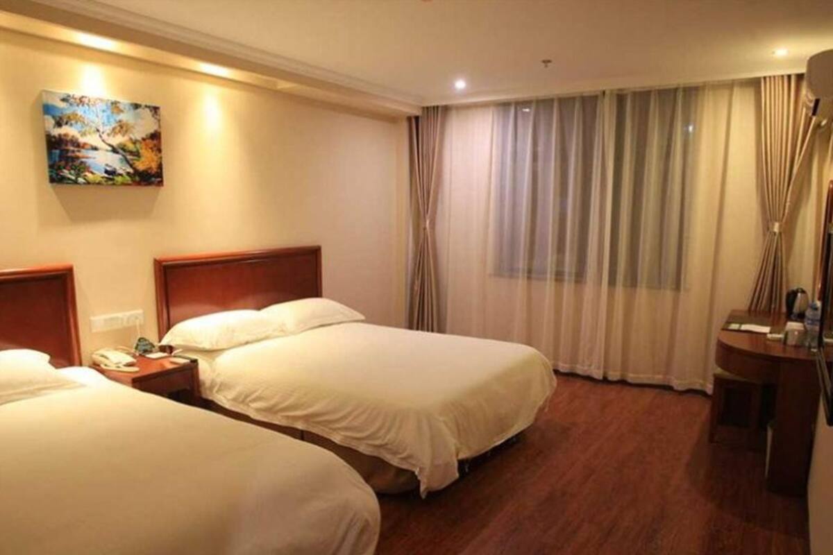 A hotel room with two beds, a painting on the wall, a desk with a lamp, and a large window with curtains.