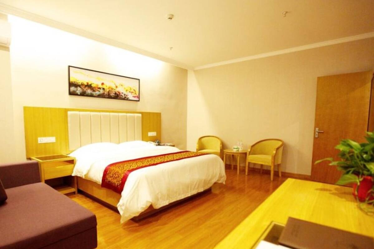 A hotel room with a bed, a bedside table, a chair, a small table with a plant, and a door.