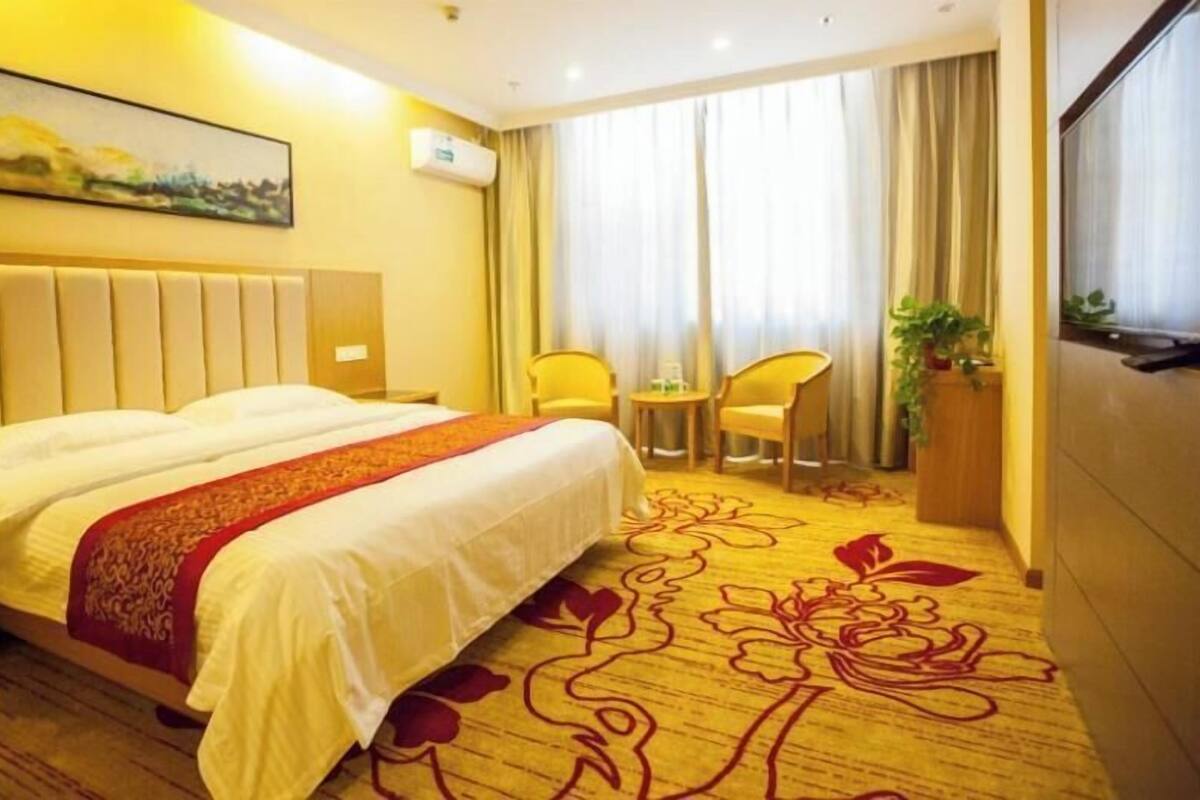 A hotel room with a large bed, two chairs, a small table, a potted plant, and a painting on the wall.