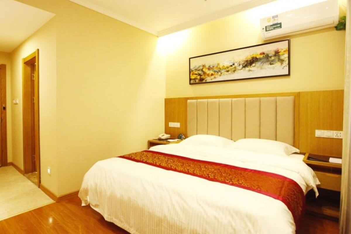A hotel room with a bed, a bedside table with a lamp, a wall-mounted air conditioner, and a framed picture on the wall.