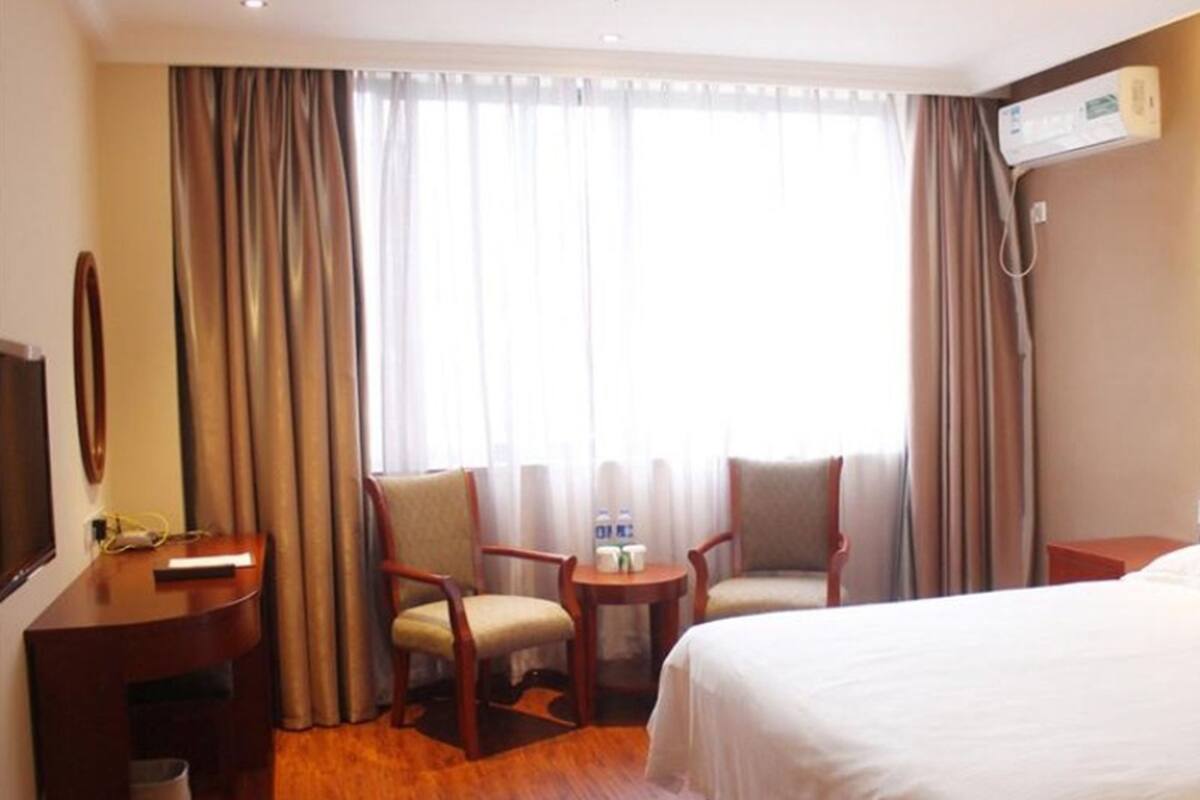 Ein Hotelzimmer mit einem Bett, zwei Sesseln, einem kleinen Tisch mit einer Wasserflasche, einem Fernseher und Vorhängen.