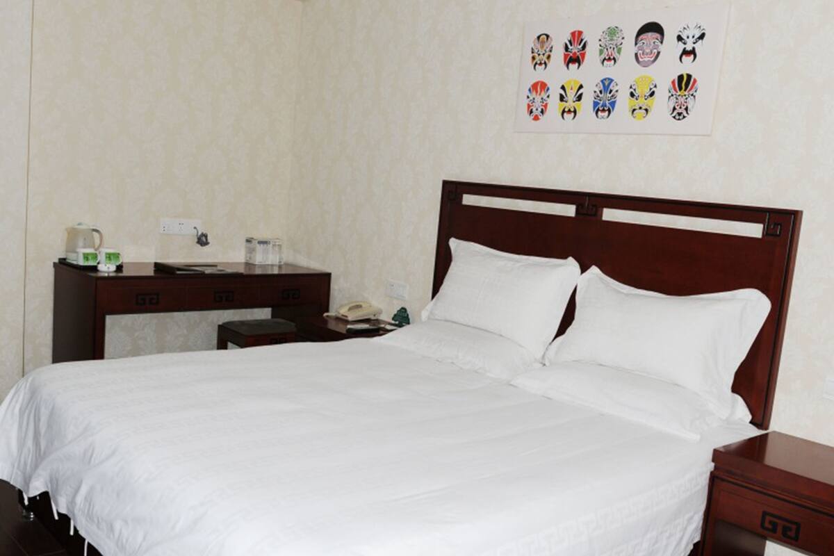 A hotel room with a large bed, a wooden nightstand, a desk with a telephone, and a wall-mounted artwork of various masks.