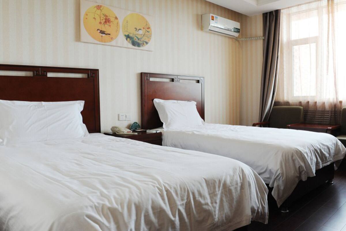 A hotel room with two single beds, a wall-mounted air conditioner, and a window with curtains.