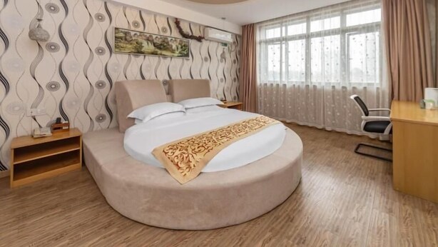 A modern hotel room with a curved bed, a desk, and a large window with curtains.