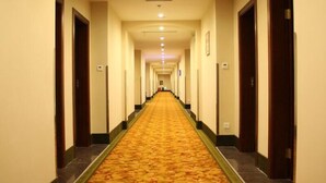 A long, narrow hotel corridor with a carpeted floor, doors on both sides, and a series of lights on the ceiling.