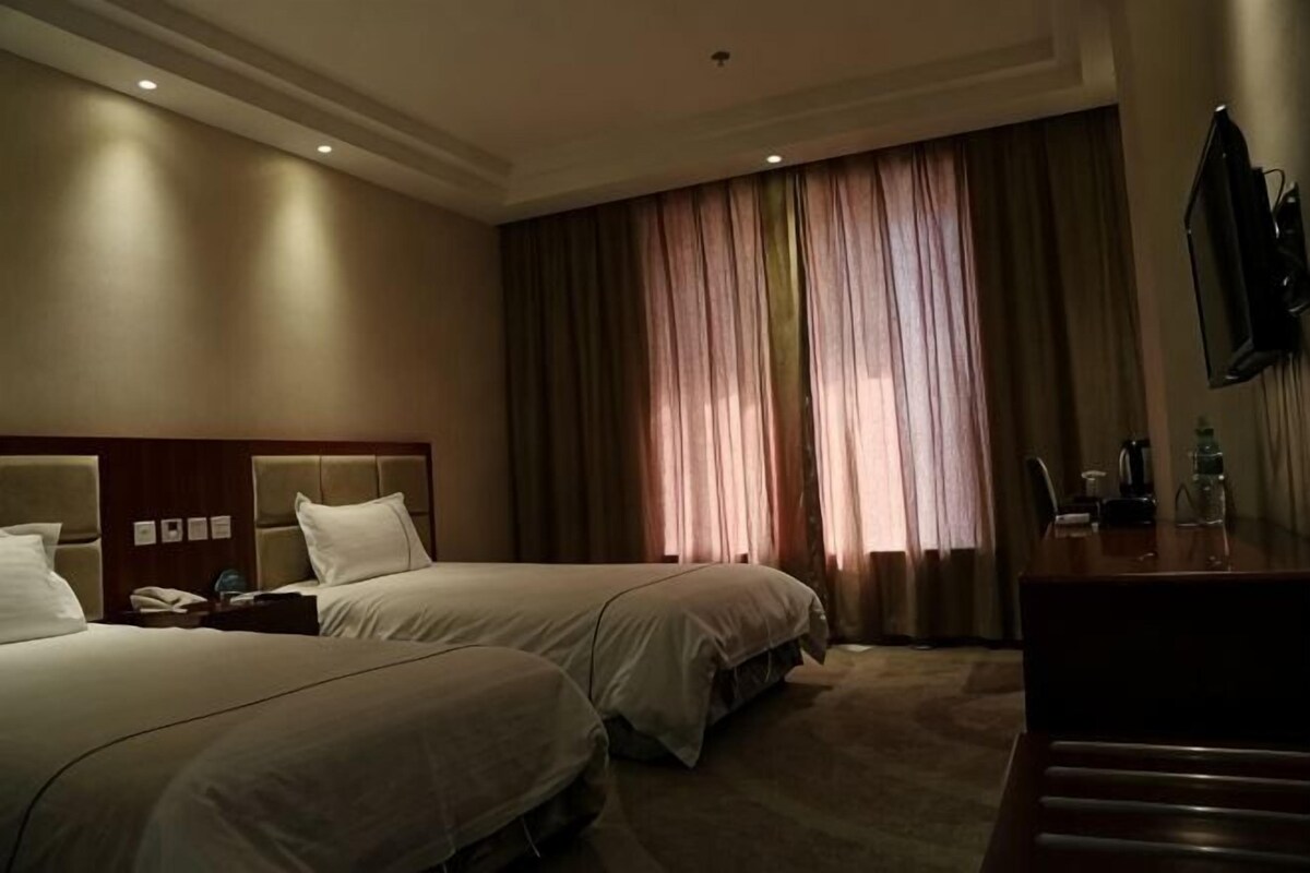 A hotel room with two beds, a desk, a TV, and a large window with curtains.