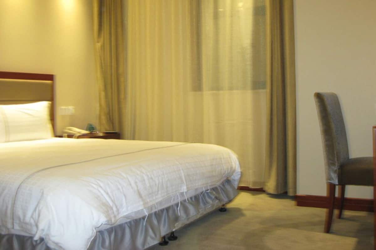A hotel room with a large bed, a chair, a bedside table with a lamp, and a window with sheer curtains.