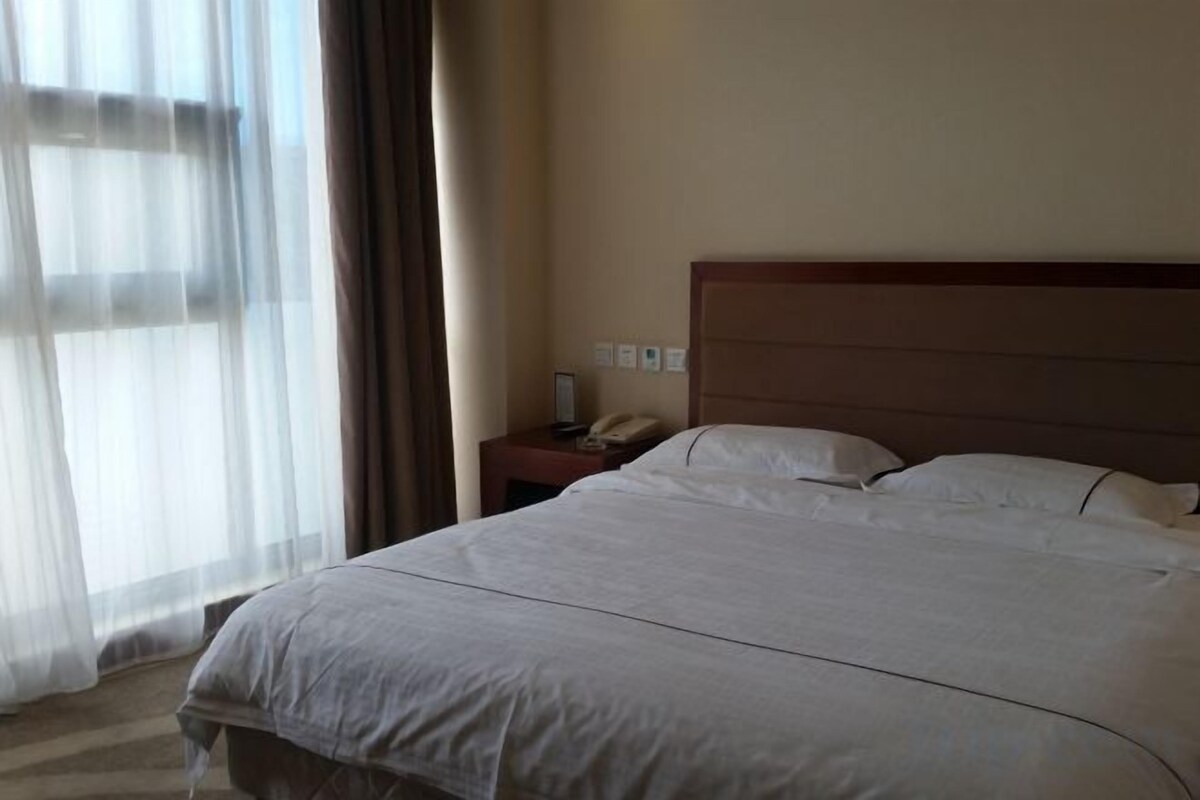 A hotel room with a large bed, a bedside table with a lamp, and a window with sheer curtains.