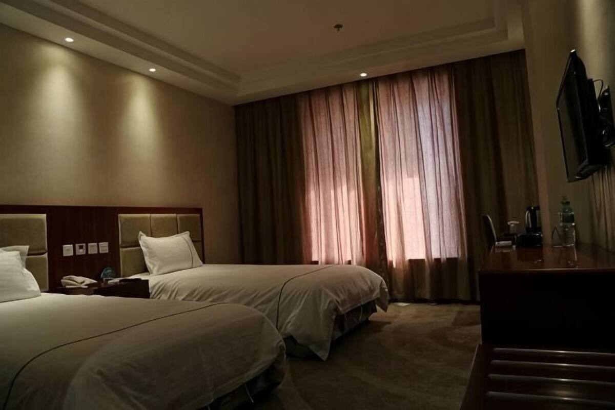 A hotel room with two beds, a desk, a TV, and a large window with curtains.