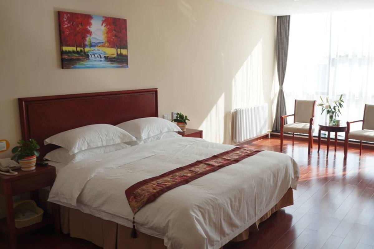 A hotel room with a large bed, a painting on the wall, a window with curtains, and a chair by the window.