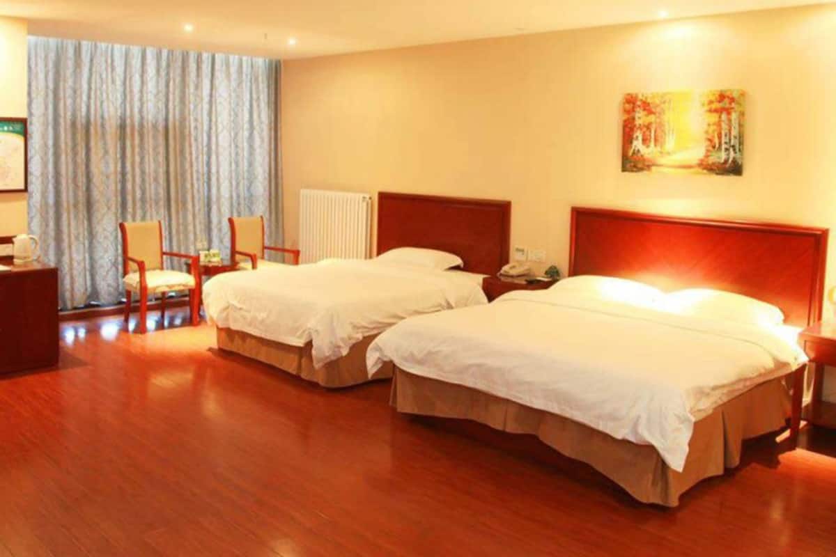 A hotel room with two beds, wooden flooring, a radiator, a painting on the wall, and a window with curtains.