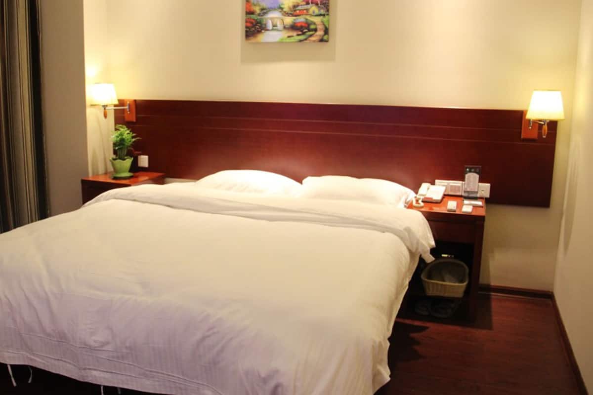 A hotel room with a large bed, two bedside tables, a nightstand, a lamp, a painting on the wall, and a small potted plant.