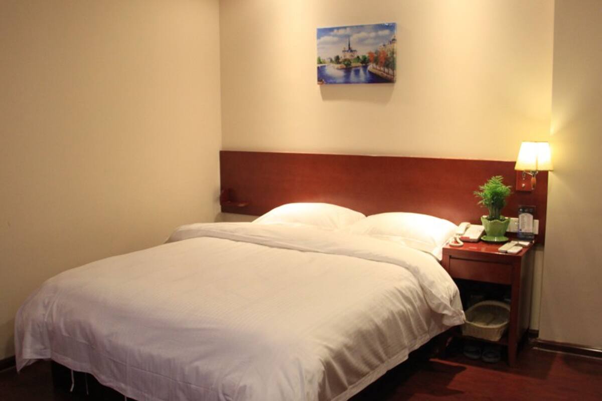 A hotel room with a large bed, a nightstand with a lamp, a painting on the wall, and a small plant.