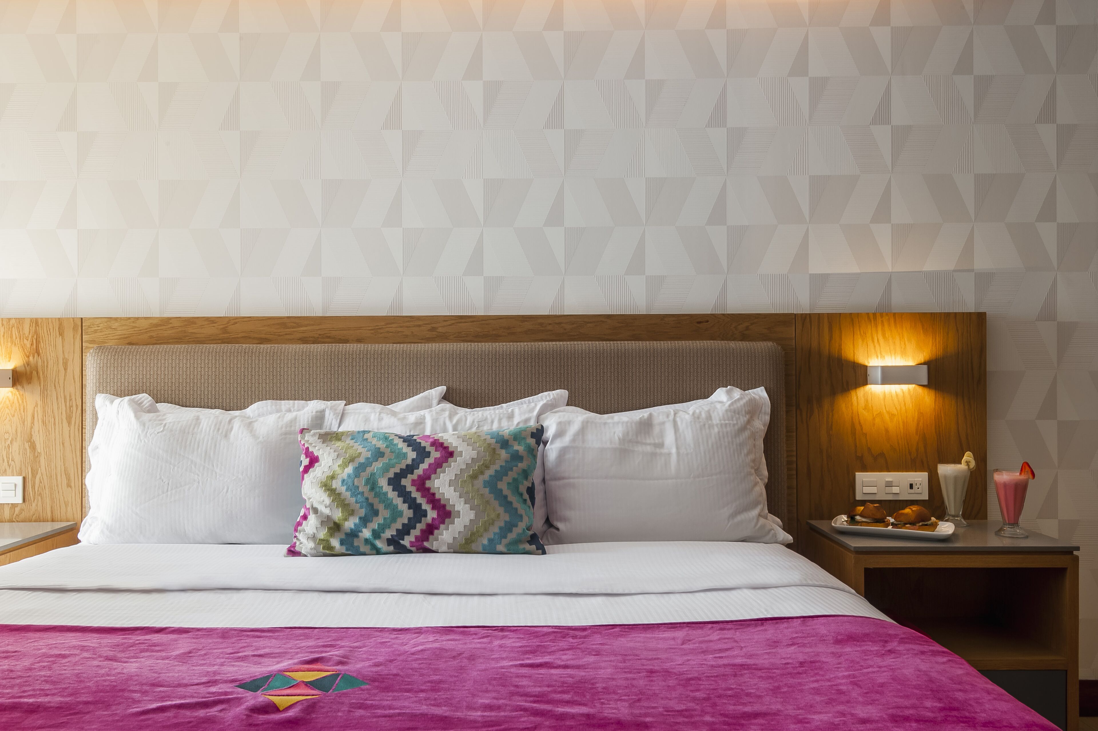 A neatly made bed with white linens and a colorful pillow, a wooden headboard, and a nightstand with a lamp and a plate of food.