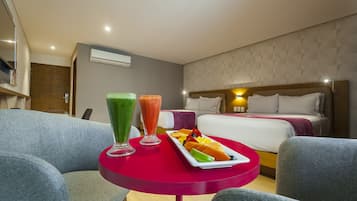 A hotel room with a bed, a red table with fruit and smoothies, and a grey sofa.