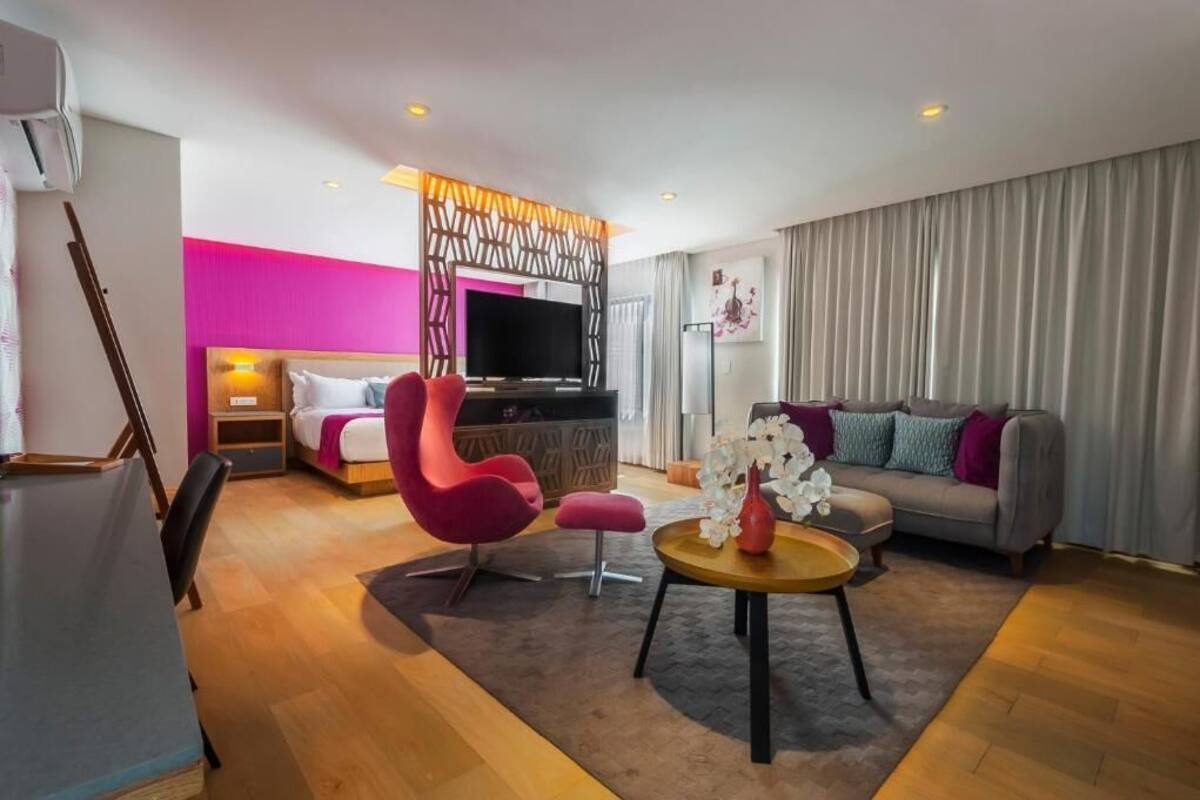 Een moderne hotelkamer met een bed, een televisie, een roze fauteuil, een bank en een klein tafeltje met bloemen.