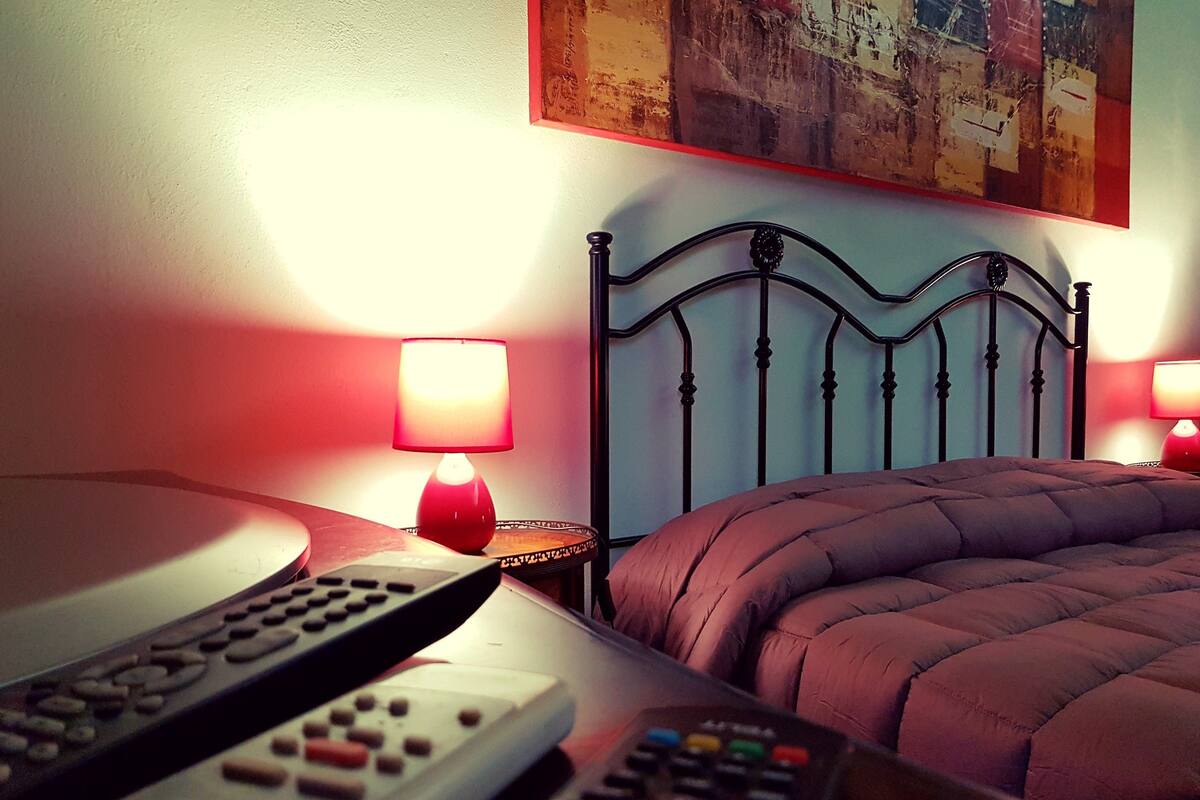A neatly made bed with a headboard, two bedside lamps, and multiple remote controls on a bedside table.