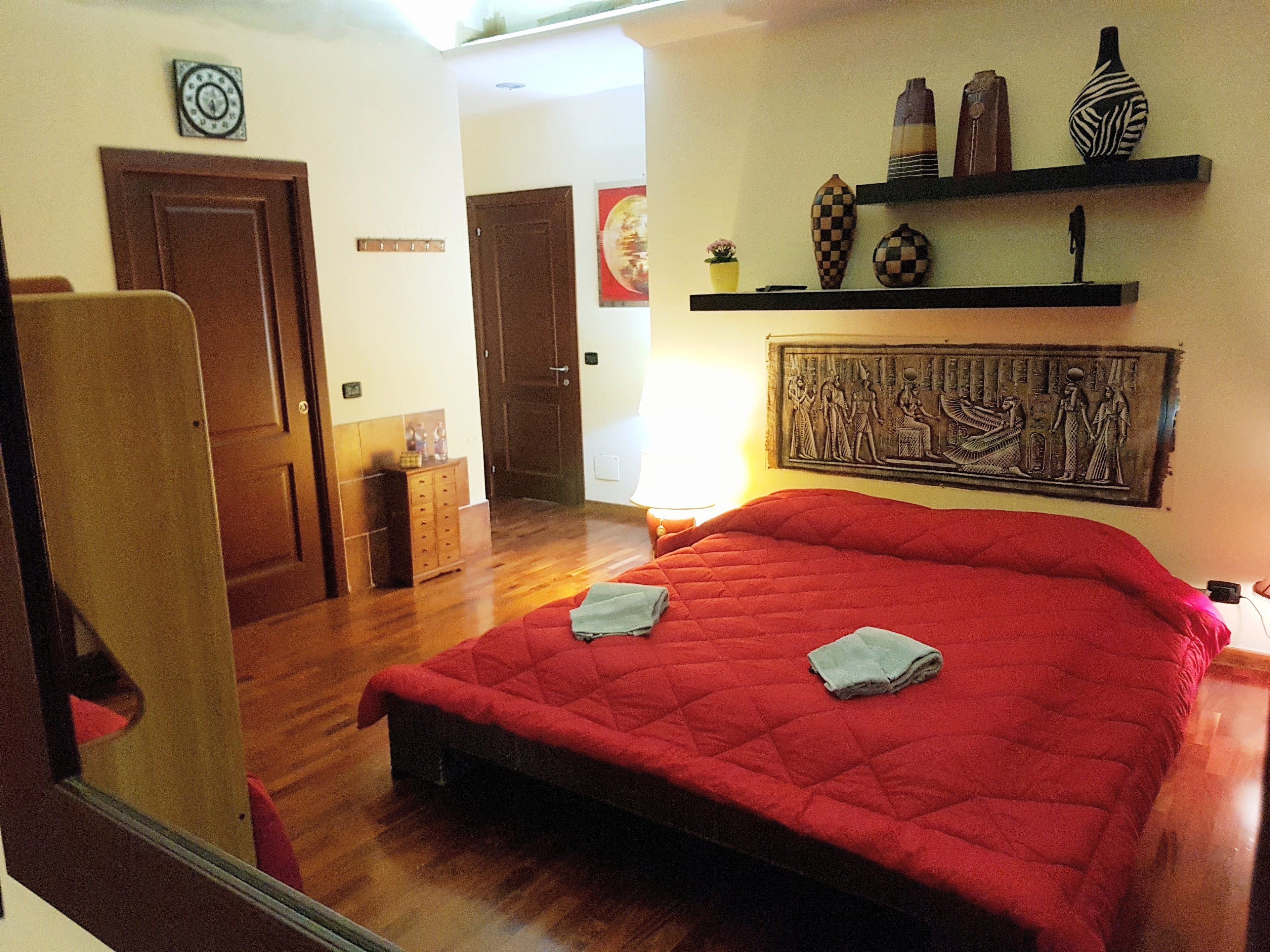 Une chambre d’hôtel avec un lit rouge, un plancher en bois, une armoire, un petit meuble, une horloge murale et des objets décoratifs disposés sur des étagères.