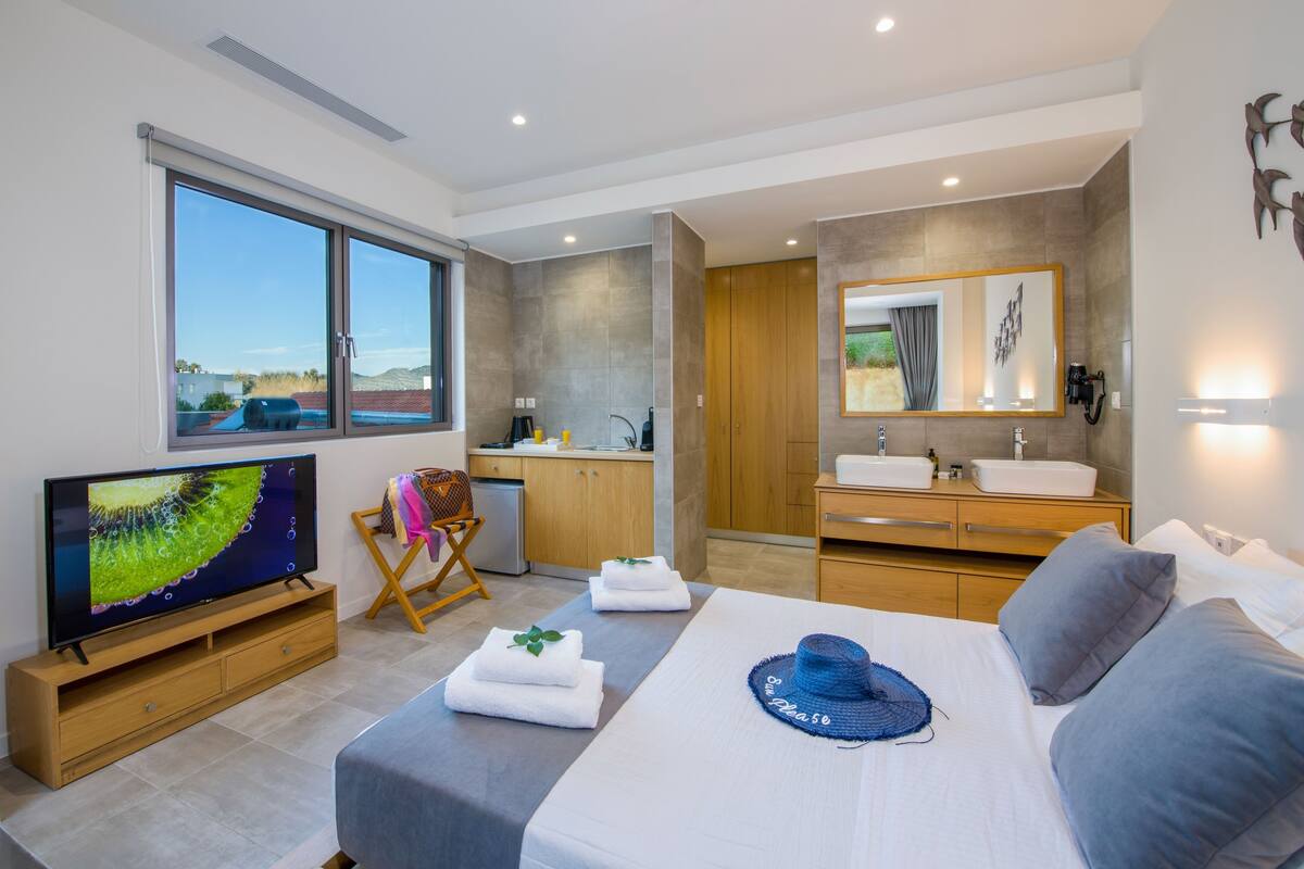 Une chambre d’hôtel moderne équipée d’une grande télévision, d’un lit avec un couvre-lit bleu, d’un bureau en bois, d’une chaise, d’un petit coin cuisine et d’une salle de bain avec un grand miroir.