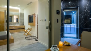 Habitación de hotel moderna con puerta de vidrio que da a un balcón, zona de comedor con mesa, cama y baño con espejo y lavamanos.
