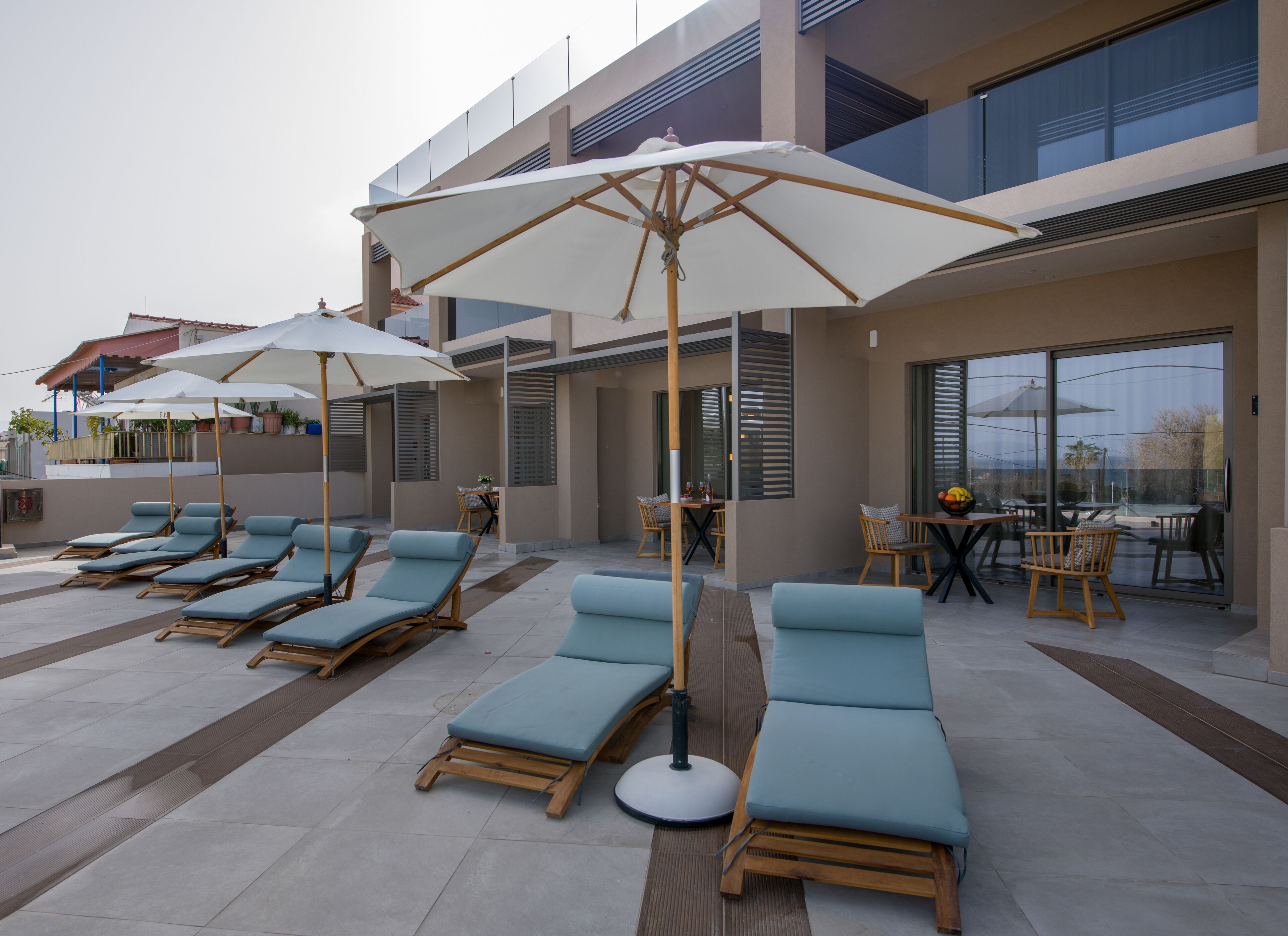 A rooftop lounge area with sun loungers and umbrellas, a dining table with chairs, and a view of the cityscape through large windows.