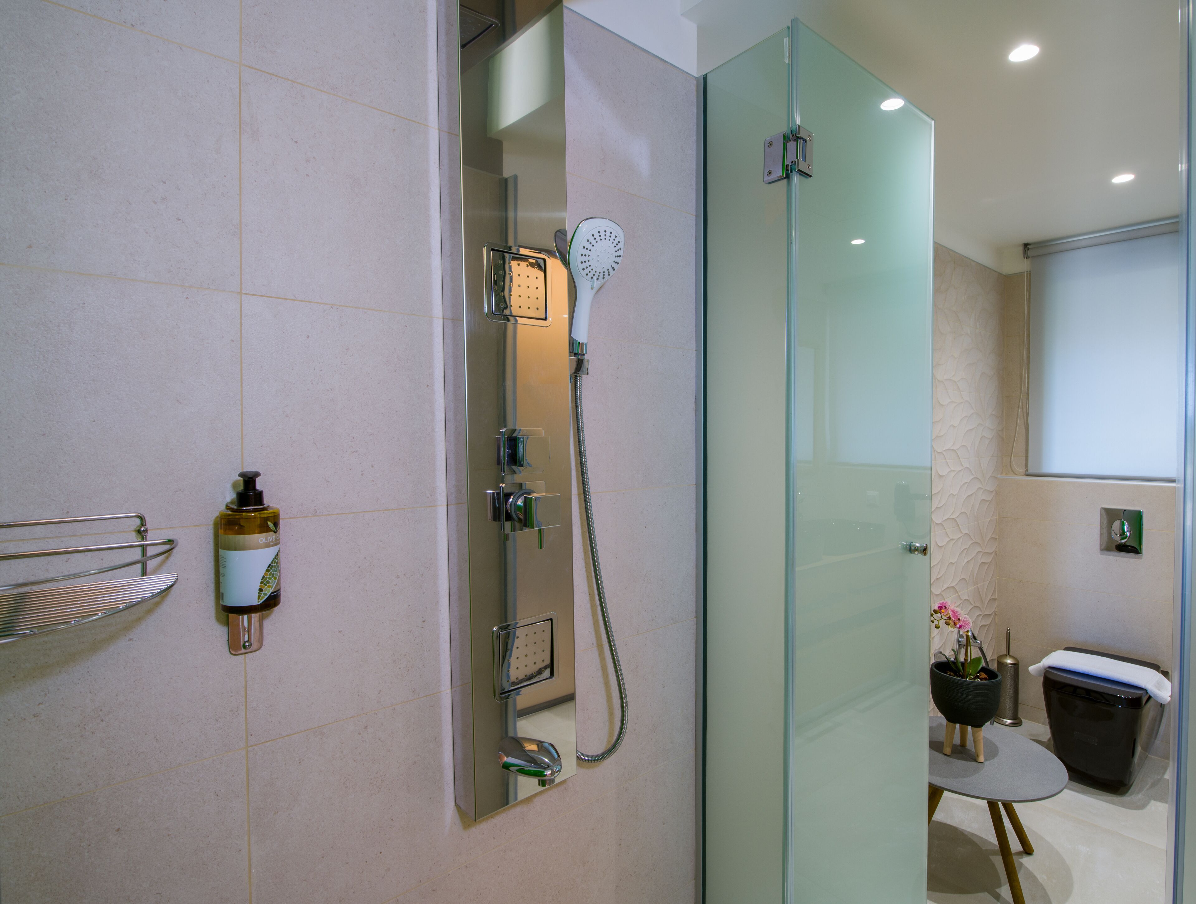 A modern bathroom with a glass-enclosed shower, a small table with a potted plant, and a trash bin.