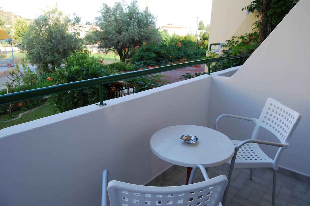 A balcony with a white table and chairs, a green railing, and a view of trees and a road.