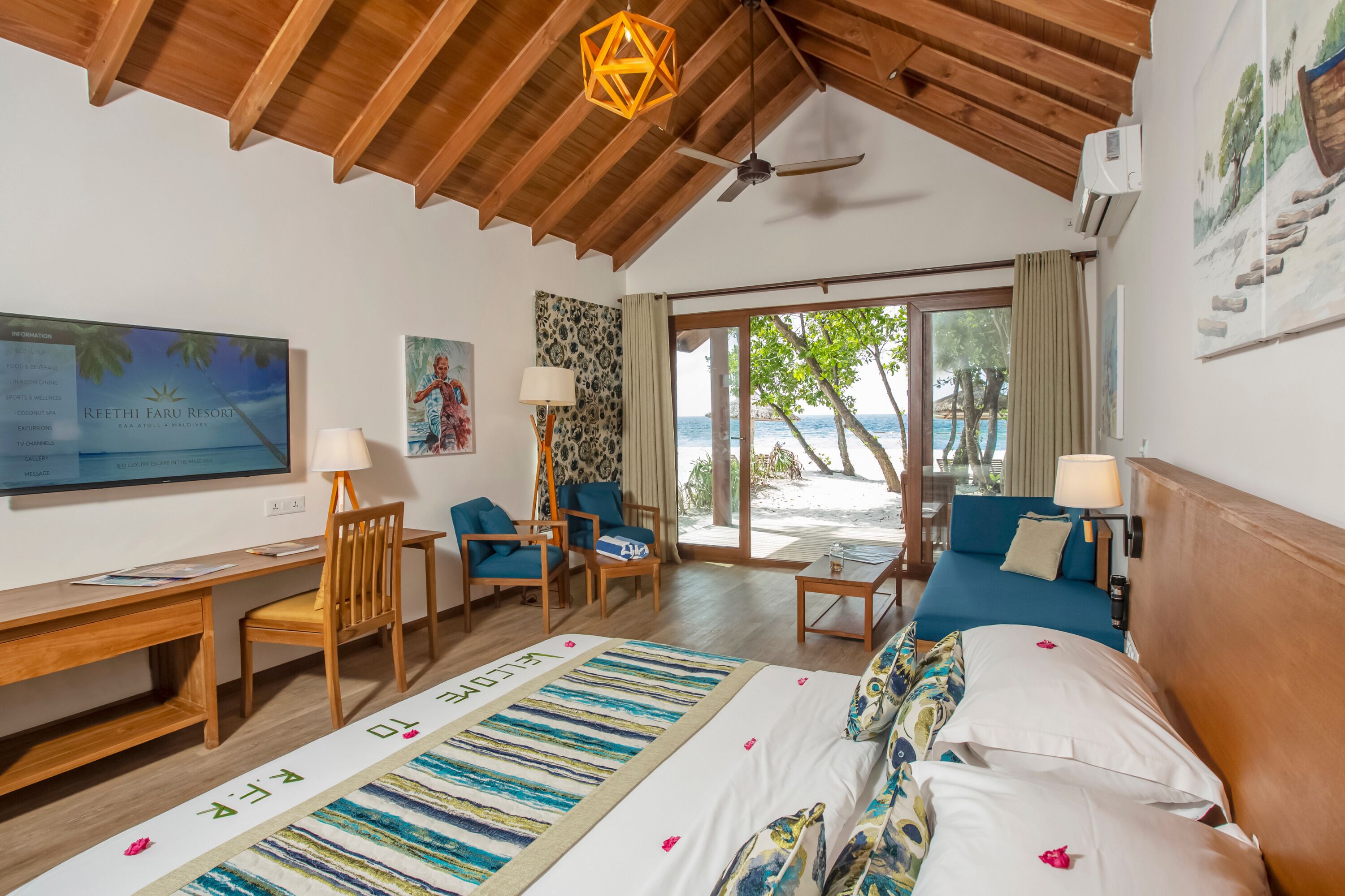 A bedroom with a wooden ceiling, a desk, chairs, a bed with a view of the beach, and artwork on the walls.