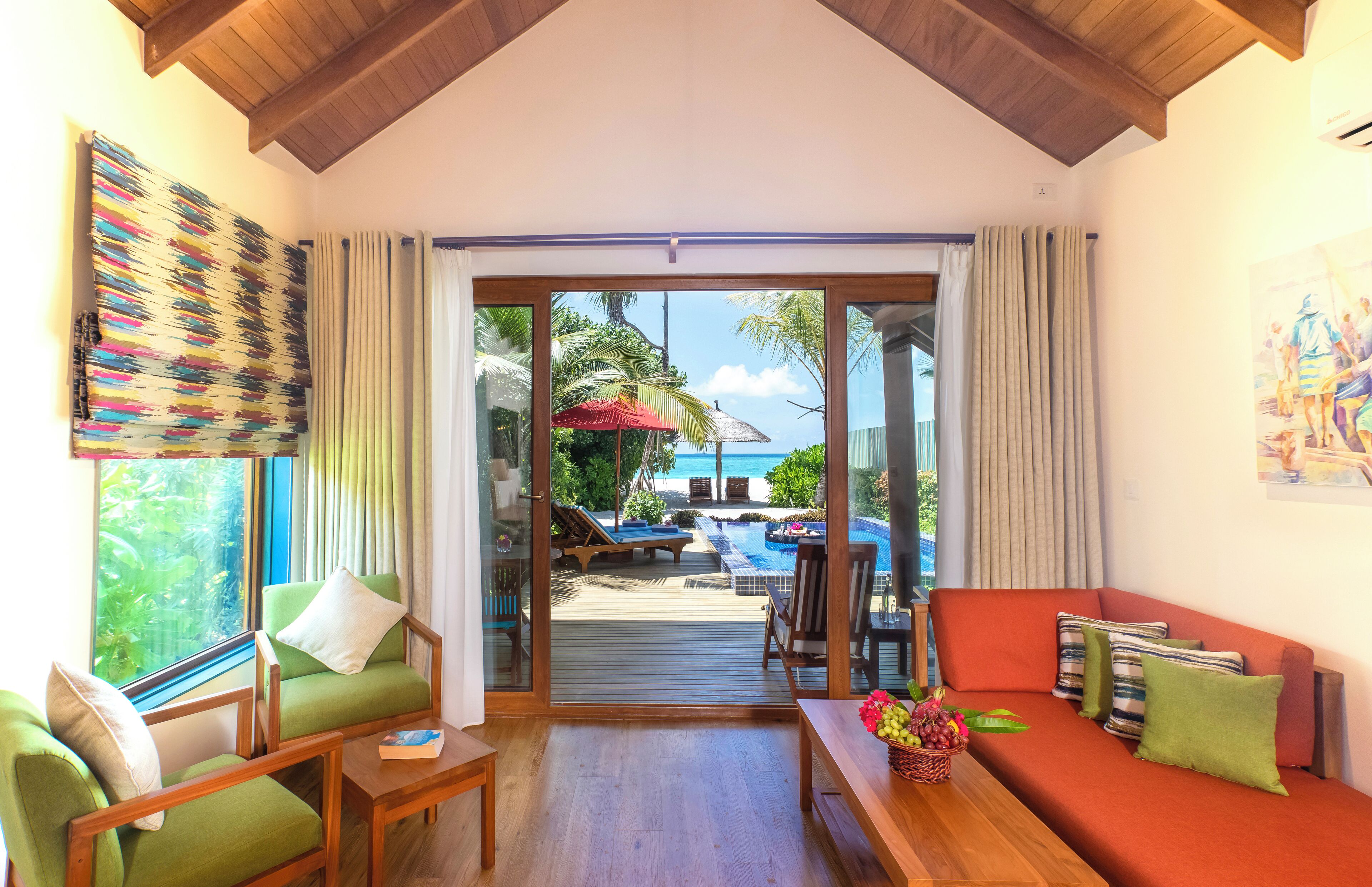 A room with a wooden ceiling, a green sofa, a red sofa, a wooden table, and a view of a pool and palm trees outside.