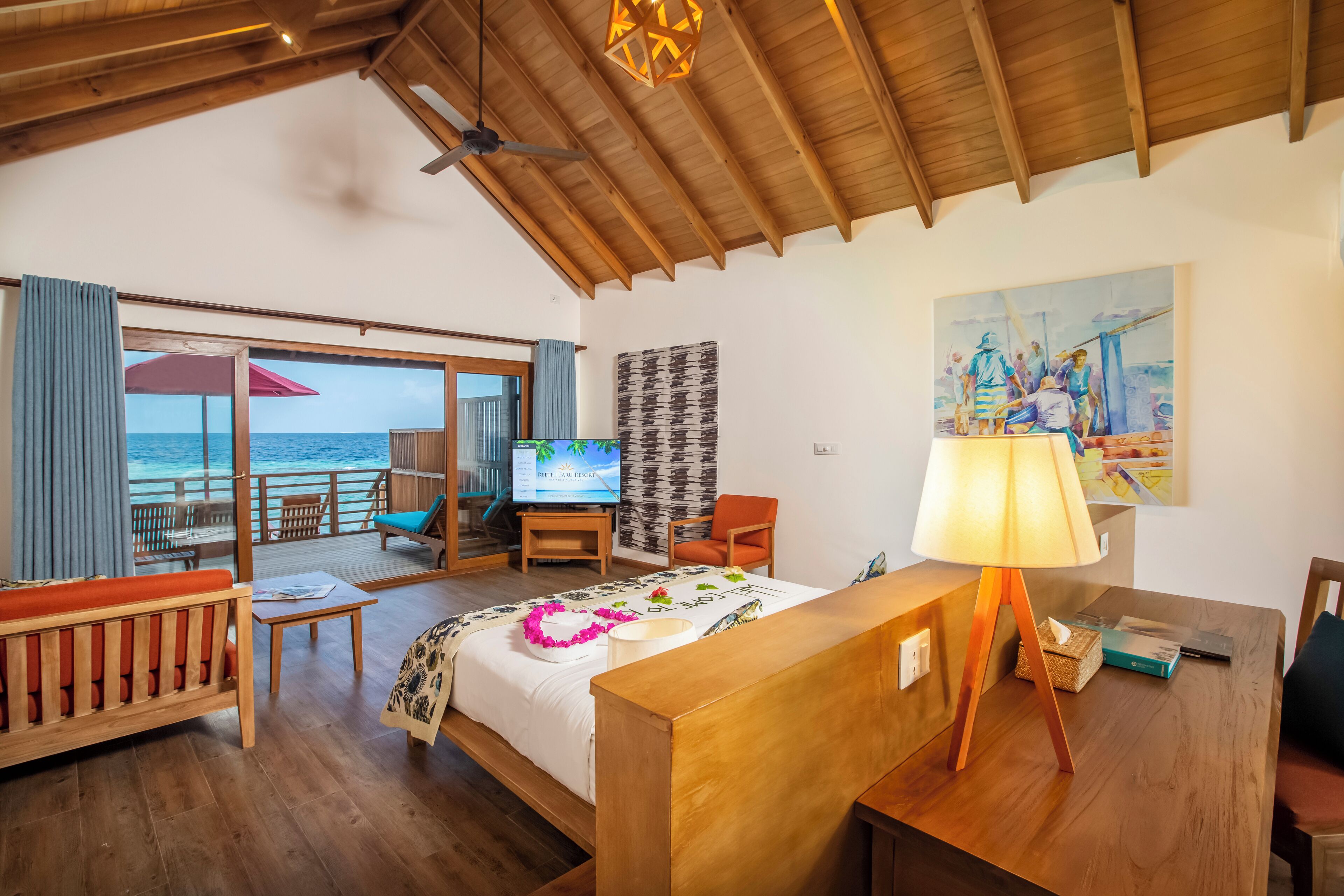 Una habitación con techo de madera, una cama con colcha estampada, un televisor, un escritorio con lámpara y un cuadro en la pared.