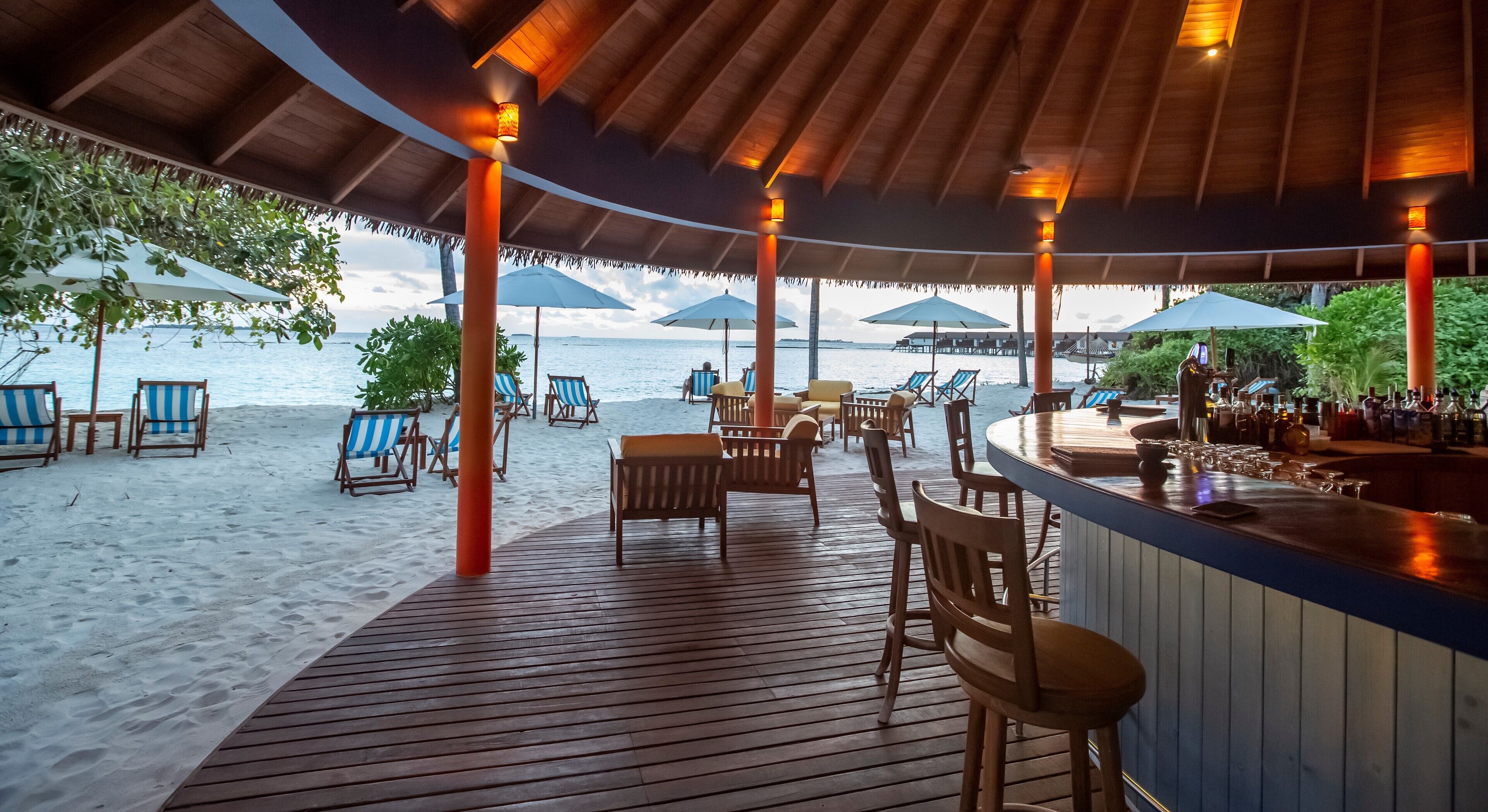 A beachside bar with wooden deck seating, umbrellas, and a view of the ocean.