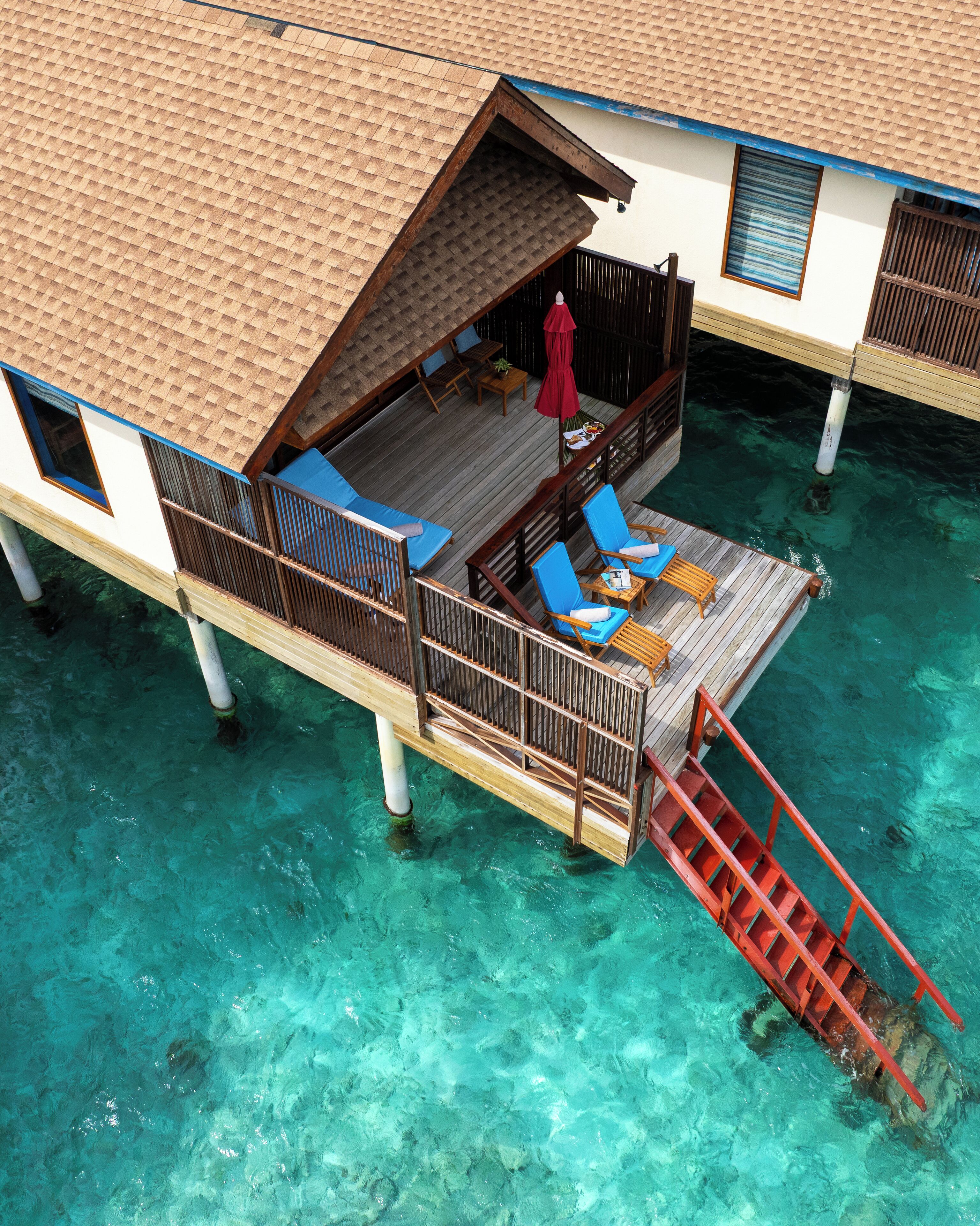 An overwater bungalow with a wooden deck, blue lounge chairs, and a red umbrella.