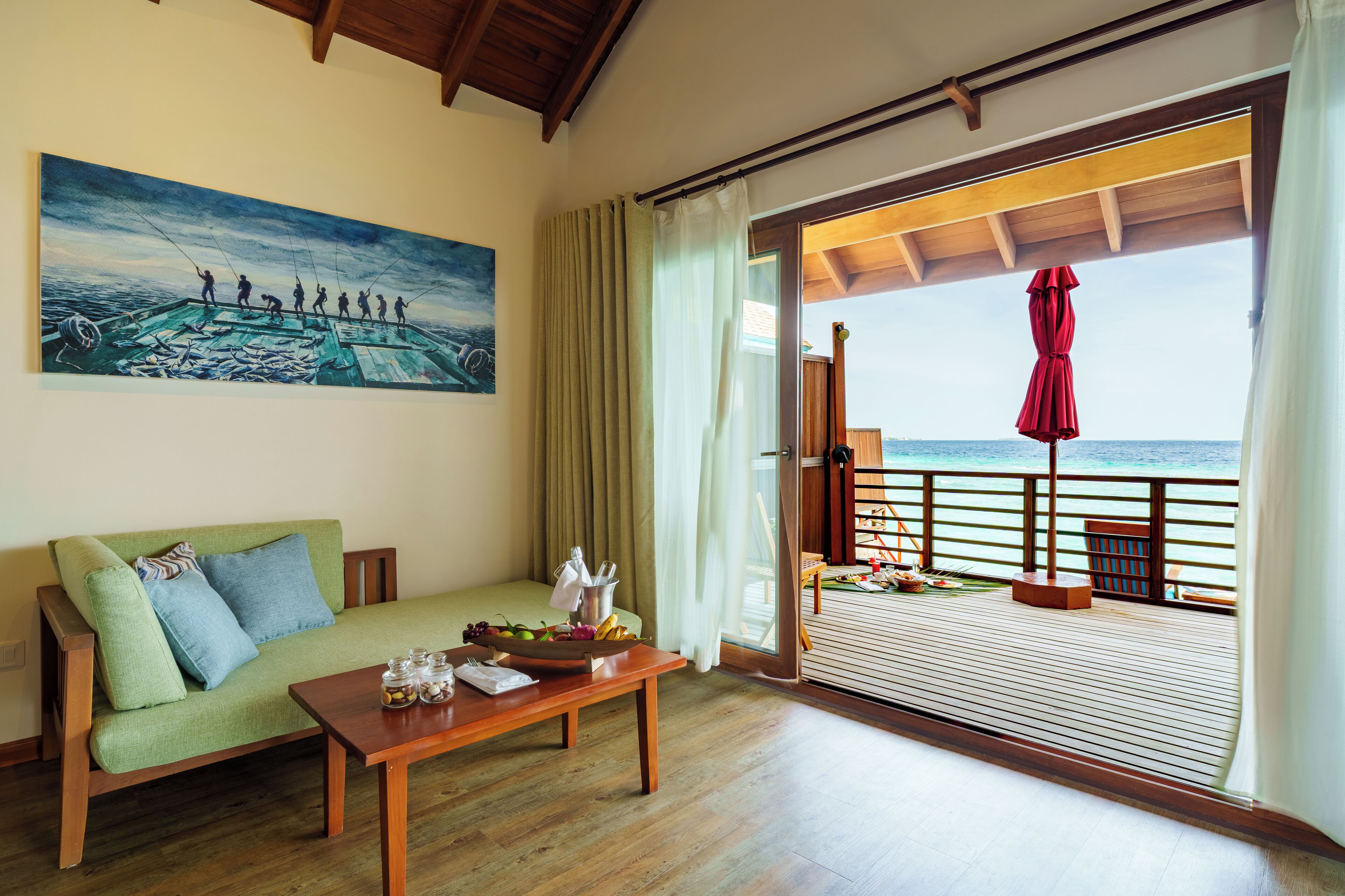 A room with a wooden floor, a green sofa with blue cushions, a wooden table with fruit and a vase, a painting of fishermen on a boat, and a balcony with a red umbrella overlooking the sea.