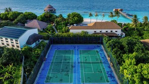 A tennis court surrounded by lush greenery, overlooking a beach with a pier and palm trees.
