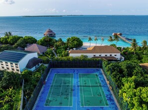 A tennis court surrounded by lush greenery, overlooking a beach with a pier and palm trees.