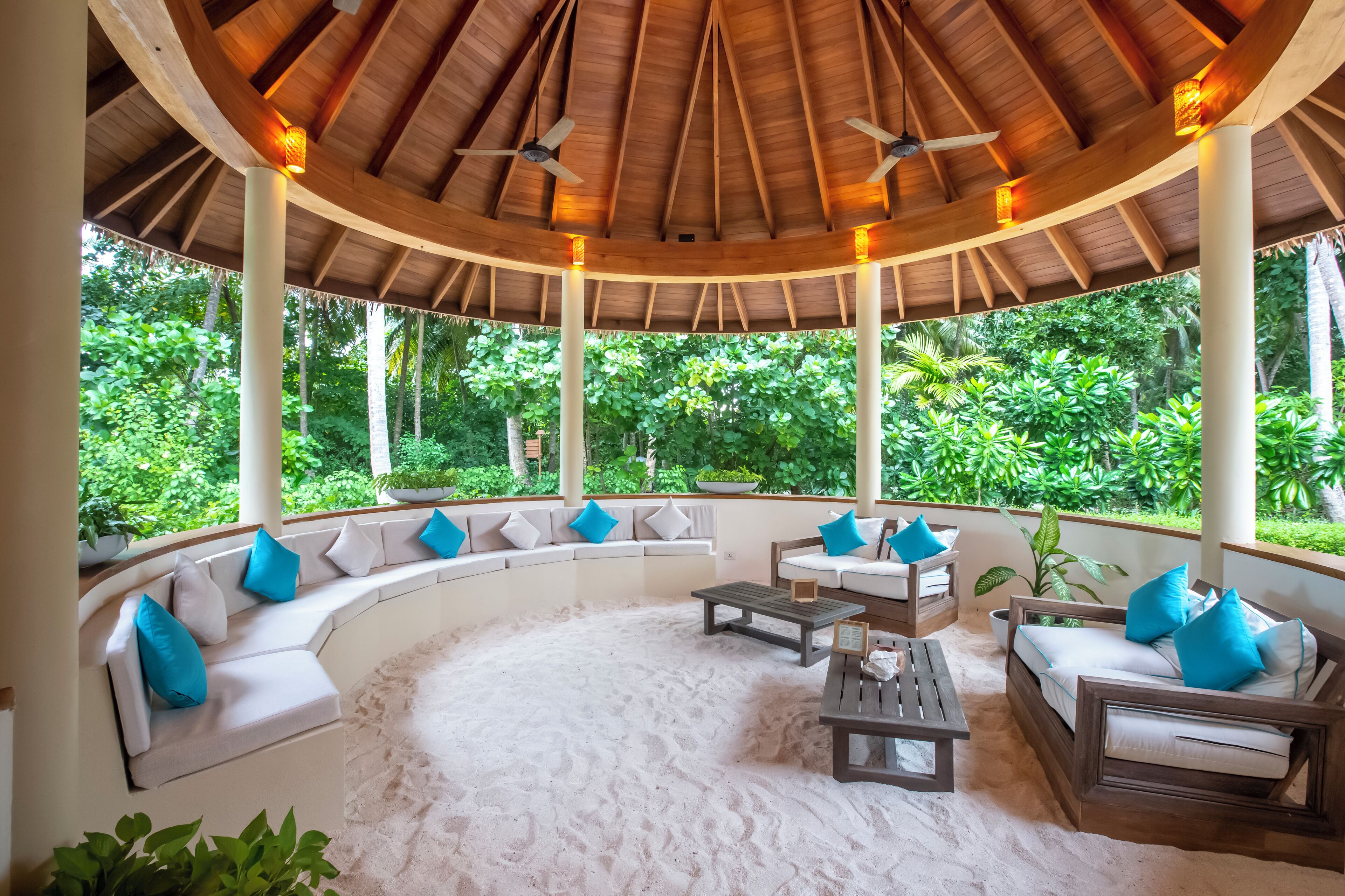 Un espacio de estar al aire libre cubierto con un banco de madera curvo, sofás blancos con cojines azules y suelo de arena.
