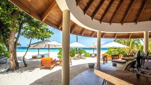 Un'area relax sulla spiaggia con pilastri in legno, poltrone da lounge, ombrelloni e un angolo bar.