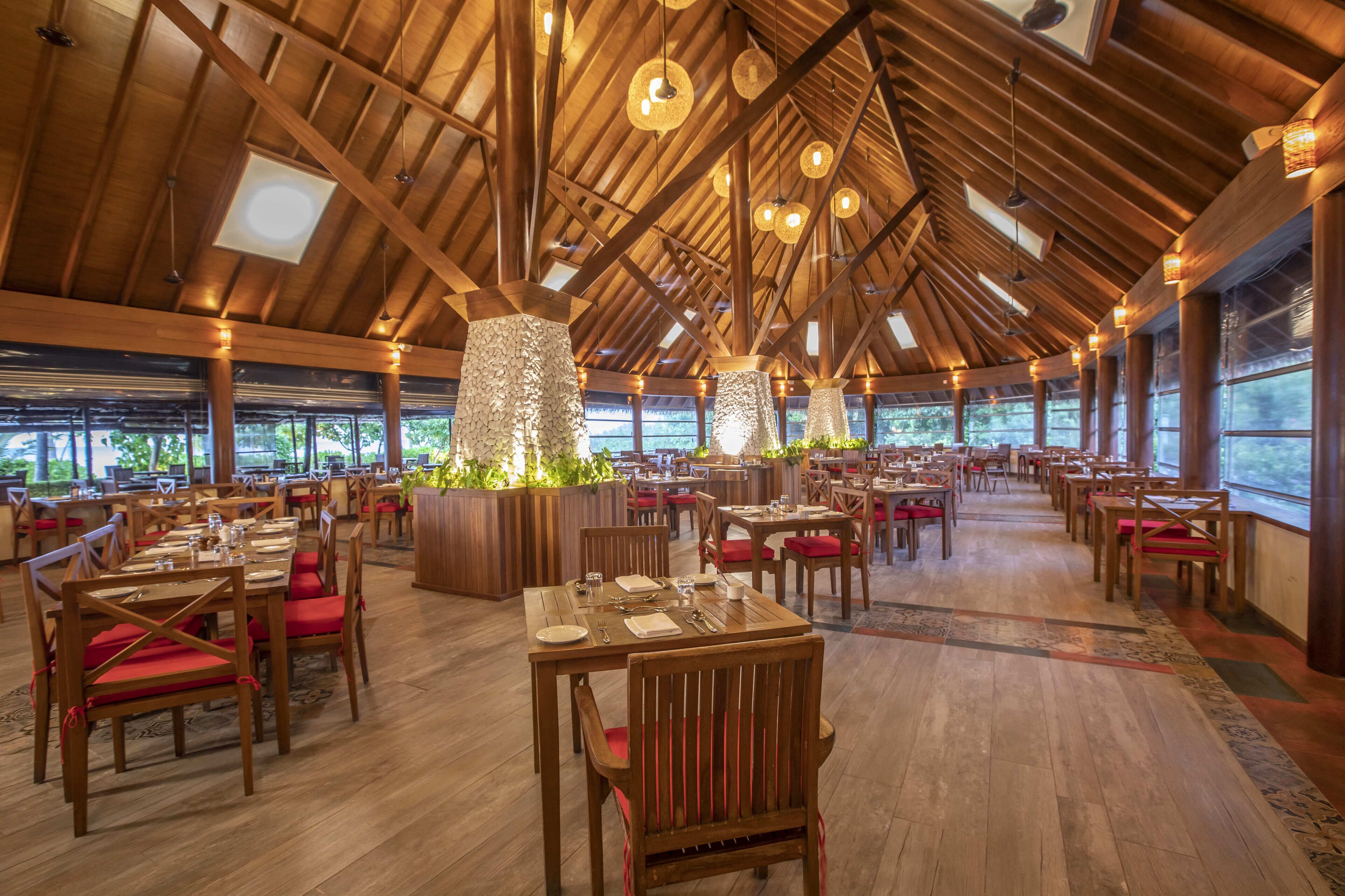 Ein geräumiges Restaurant mit Holztischen und -stühlen, großen Fenstern und einer hohen Holzbalkendecke mit hängenden Lampen.