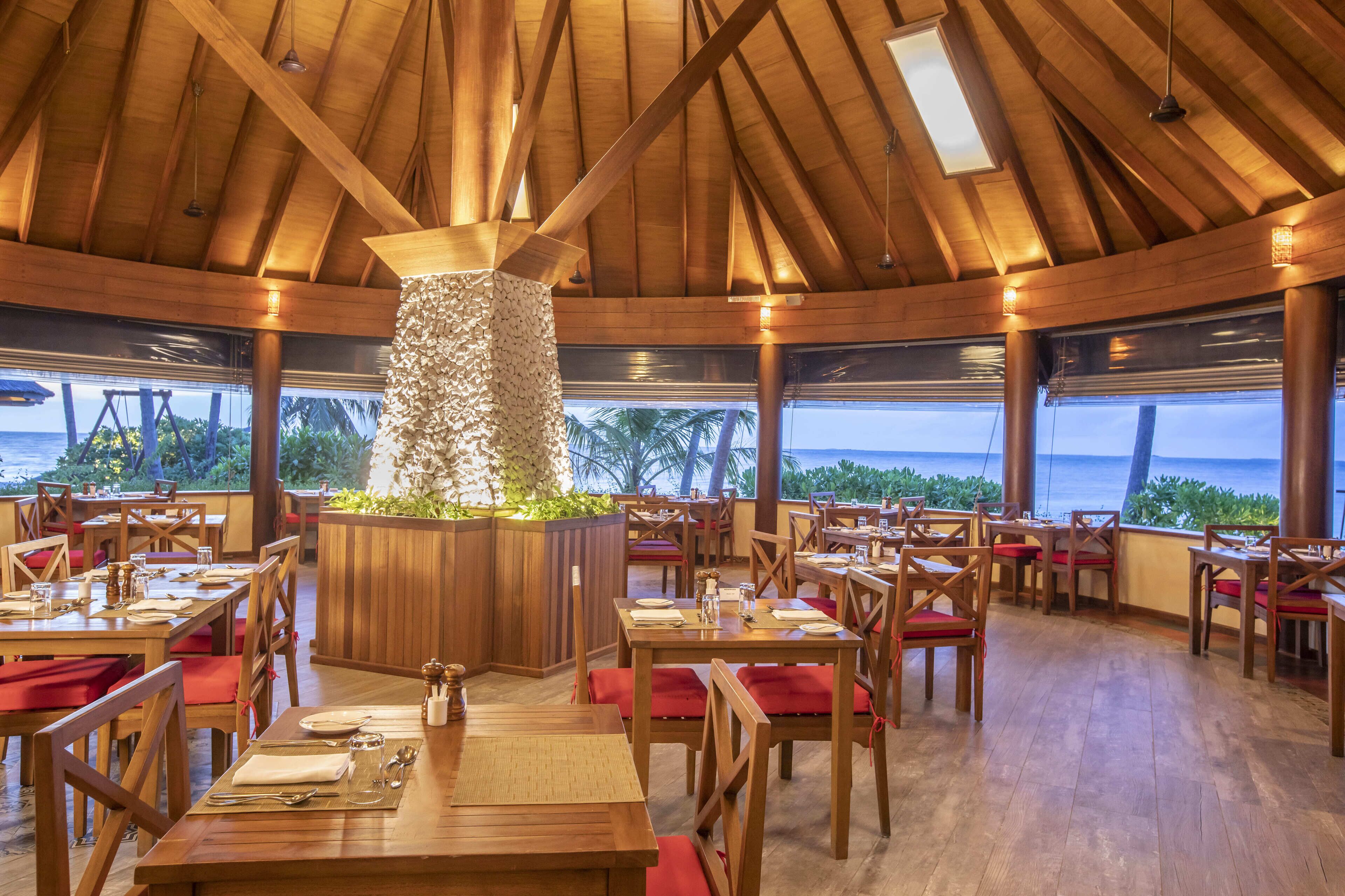 Un restaurant avec des tables et des chaises en bois, un pilier décoratif central et une vue sur l’océan.