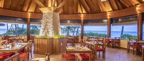 A restaurant with wooden tables and chairs, a central decorative pillar, and a view of the ocean.