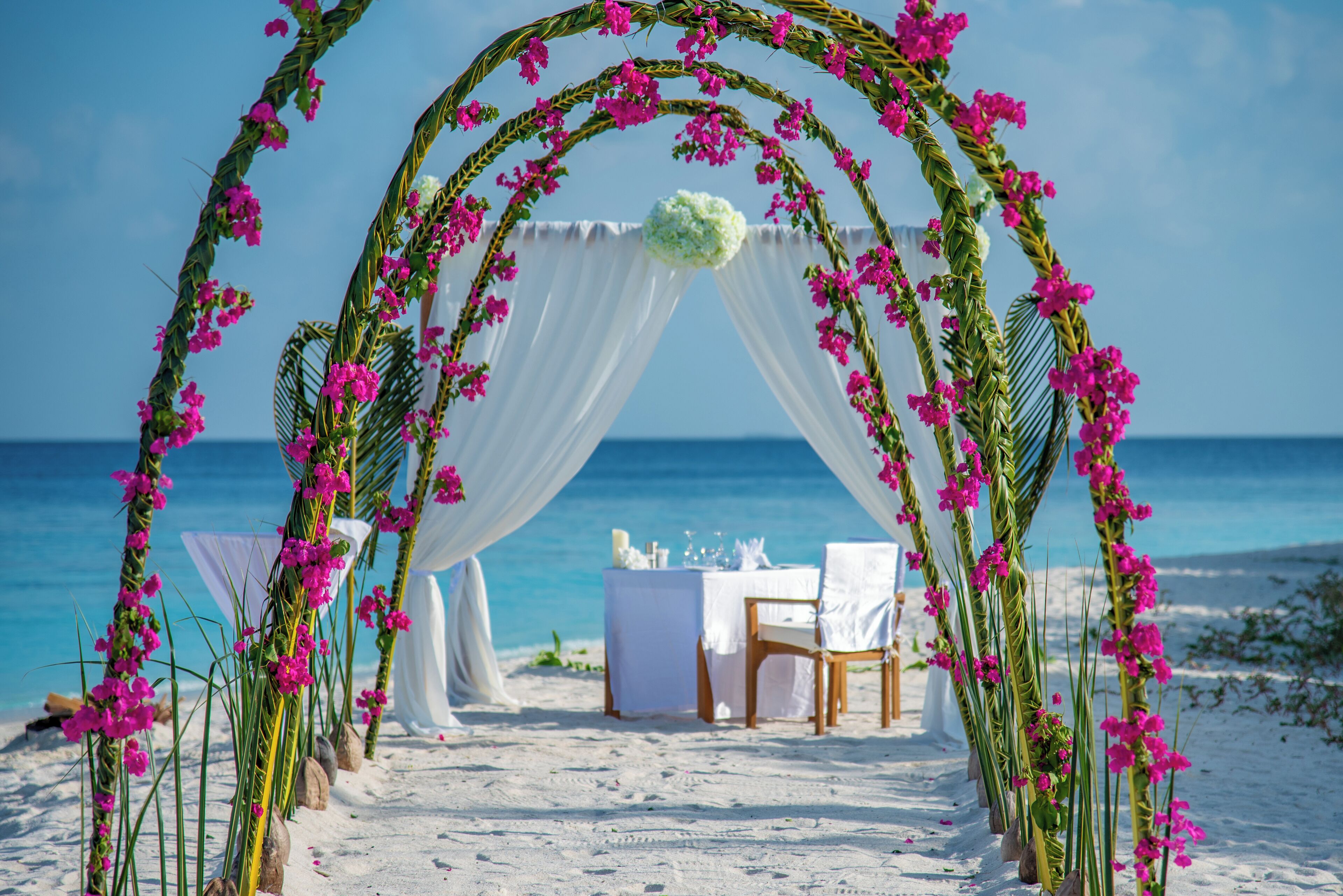 A beach wedding setup with a white arch decorated with pink flowers and a table set for a ceremony.